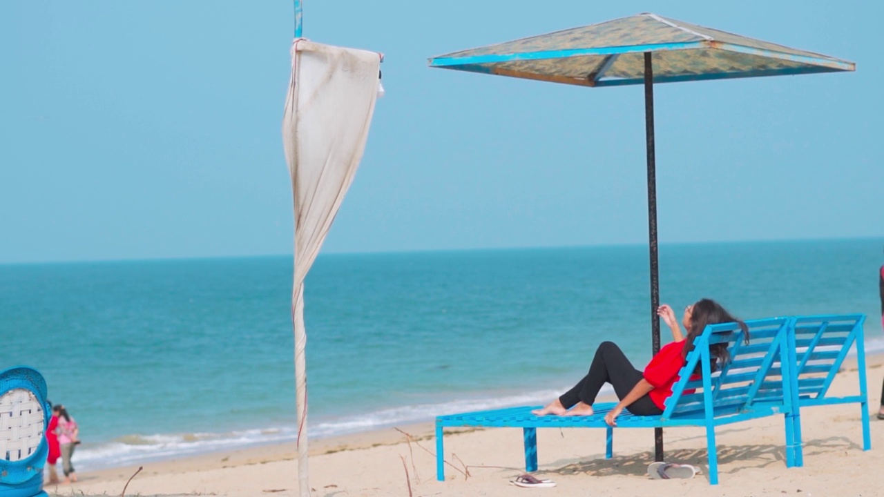 侧面的印度女人放松在甲板椅子下的海滩伞在前面的大海。在印度古吉拉特邦的Shivrajpur海滩。暑假与旅游的概念。海滩度假胜地的背景视频素材