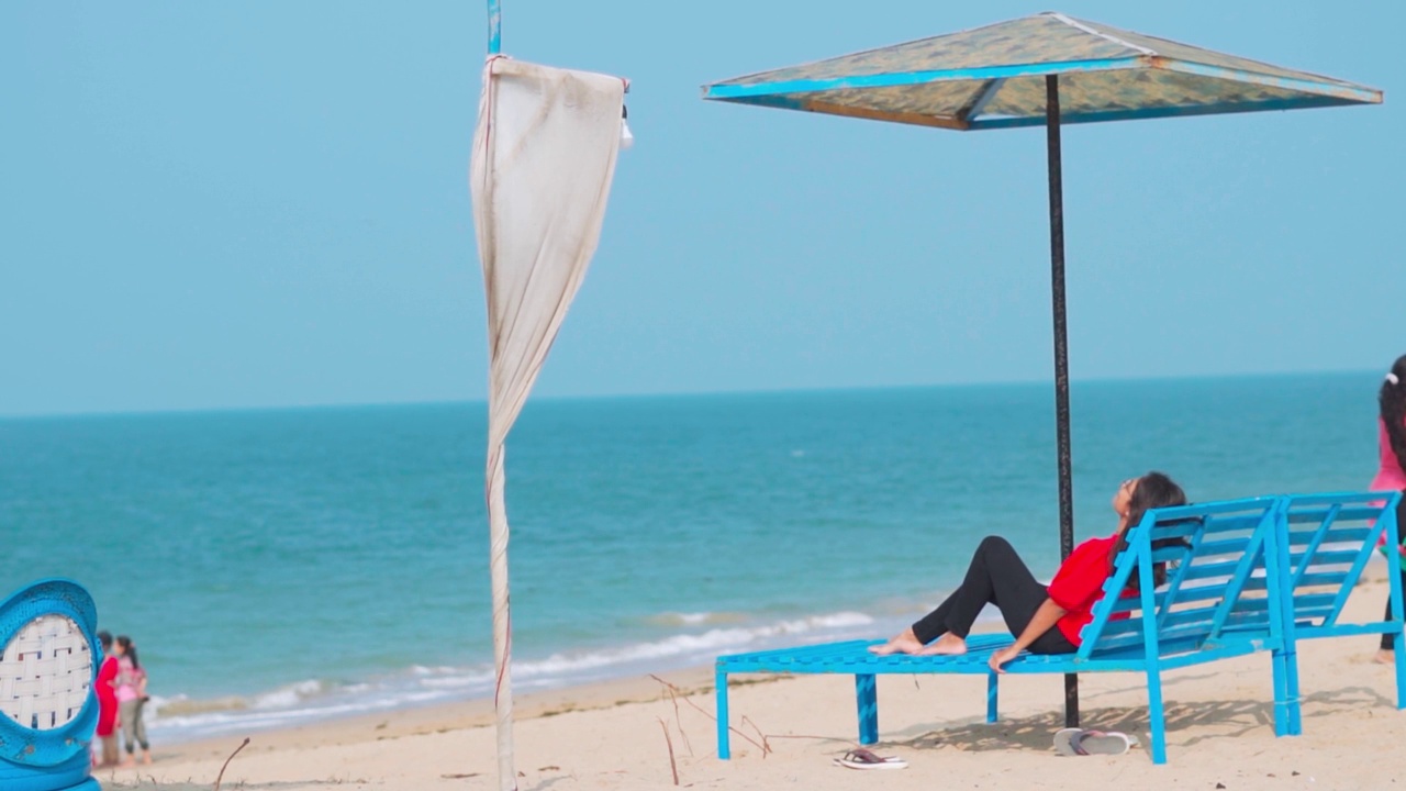 侧面的印度女人放松在甲板椅子下的海滩伞在前面的大海。在印度古吉拉特邦的Shivrajpur海滩。暑假与旅游的概念。海滩度假胜地的背景视频素材