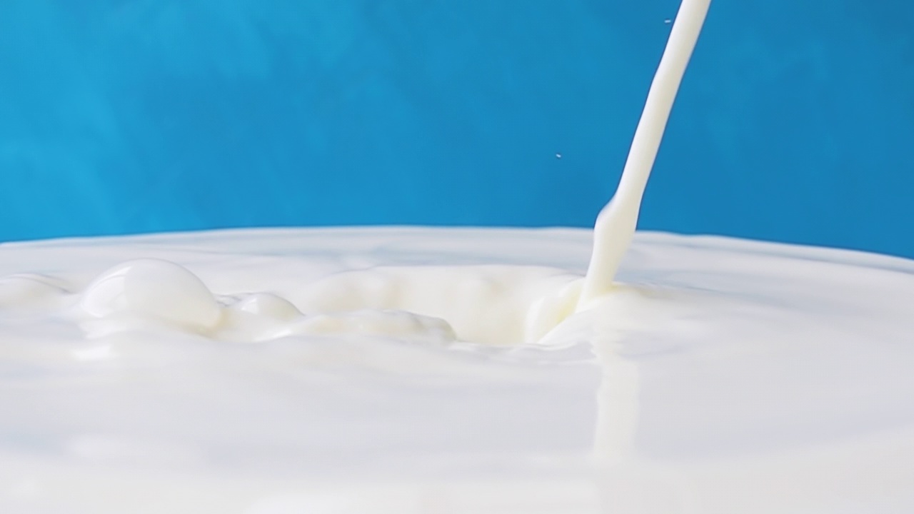 牛奶倒在牛奶表面的特写与美丽的飞溅和滴在蓝色的背景。4k原始慢动作视频。用高速电影摄影机拍摄，1000帧/秒。视频素材