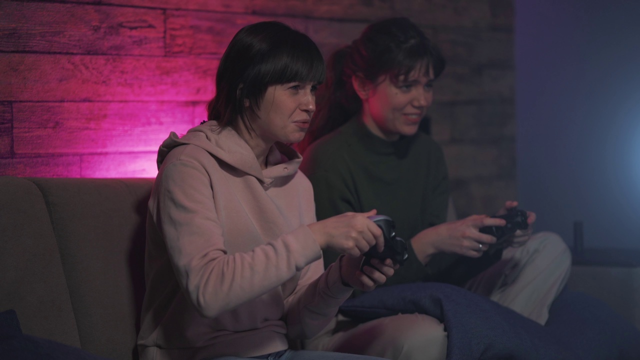 两个年轻的女人坐在沙发上用手柄玩游戏机。女性玩家在家一起享受快乐和微笑的在线娱乐视频素材
