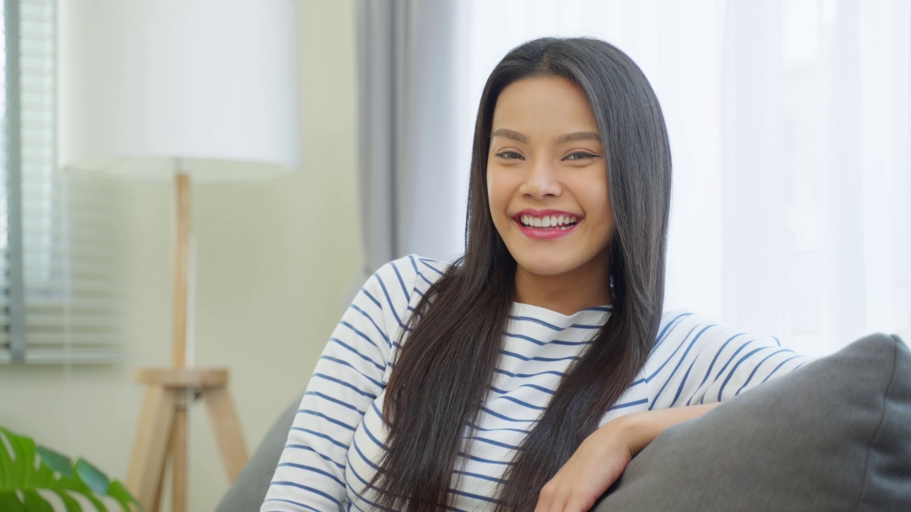 亚洲美女的肖像坐在家里客厅的沙发上。有吸引力的年轻女性感觉快乐和放松享受清晨周末活动的生活方式在房子和微笑，看着相机。视频素材