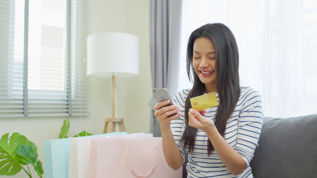 亚洲美女喜欢在家里的客厅网上购物。漂亮的女孩坐在沙发上，用手机在网上商店购买商品后拿着信用卡进行网银网上支付。视频素材