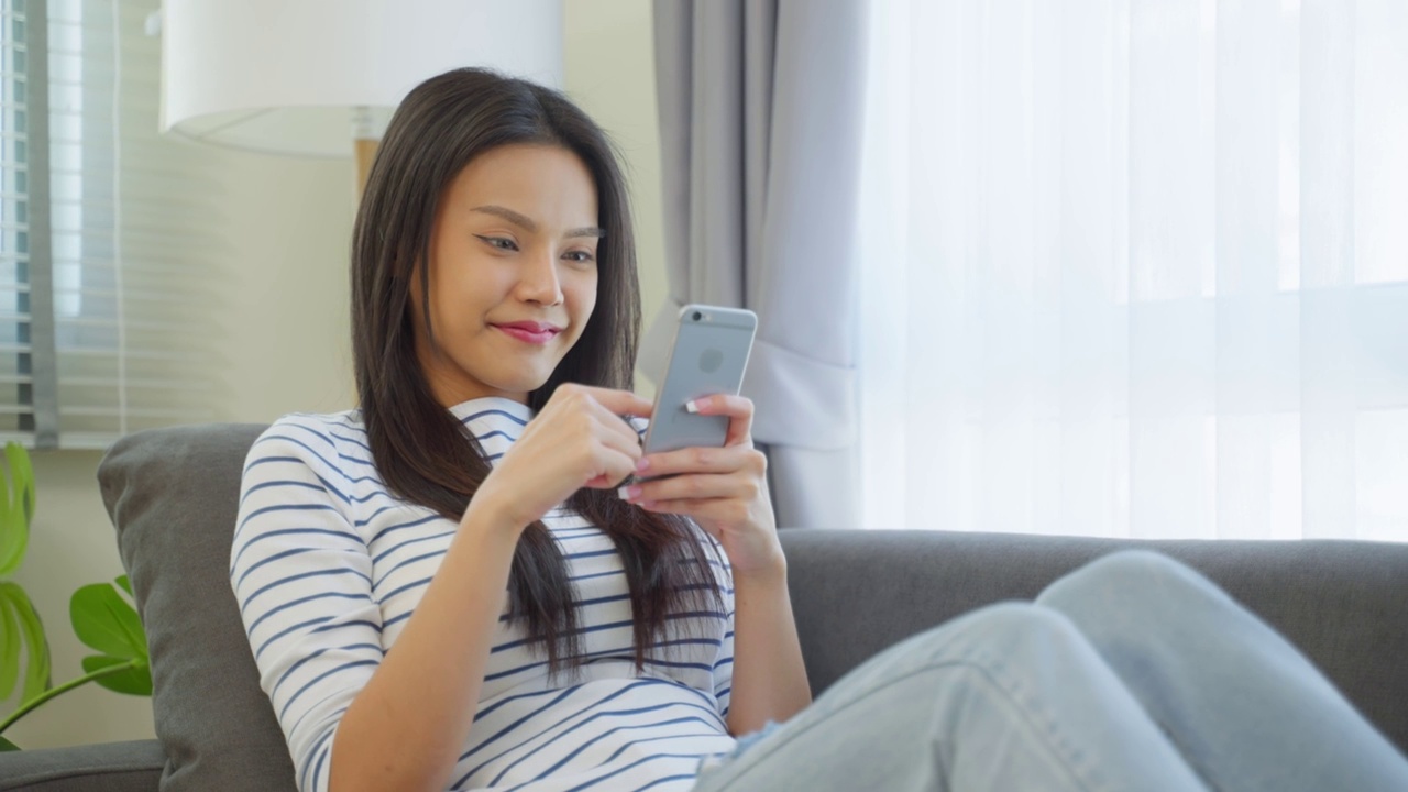 亚洲美女坐在家里的沙发上用手机聊天。快乐迷人的年轻女孩在家里度过休闲时间，感到放松，享受在客厅用智能手机交流和讨论。视频素材