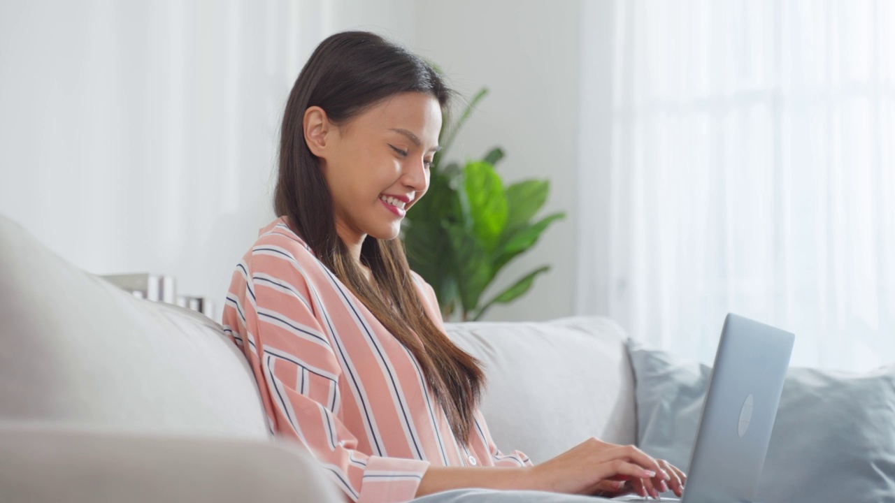 年轻的亚洲女商人在电脑上打字，在家工作。有魅力的漂亮自由职业女职工坐在沙发上，在客厅用笔记本电脑带着幸福和微笑呆在家里。视频素材