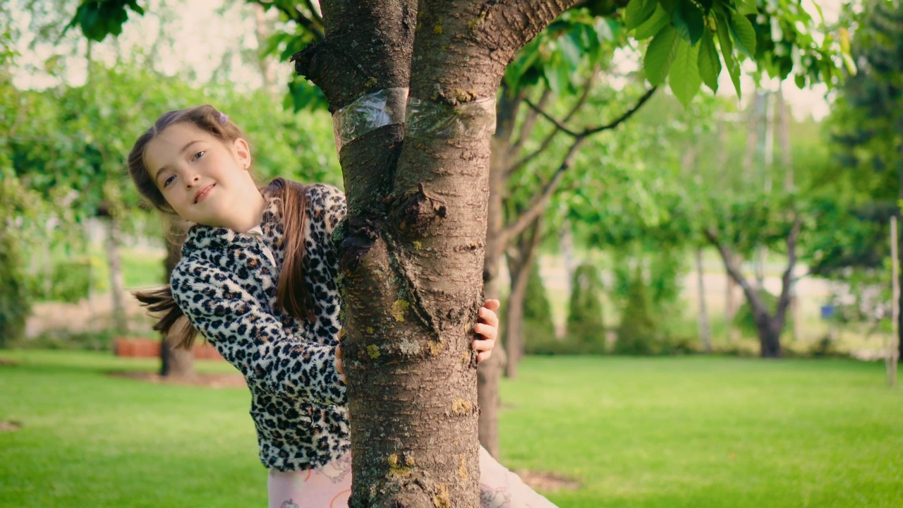 开朗微笑的女孩躲在树后，从树后探出头来笑视频素材