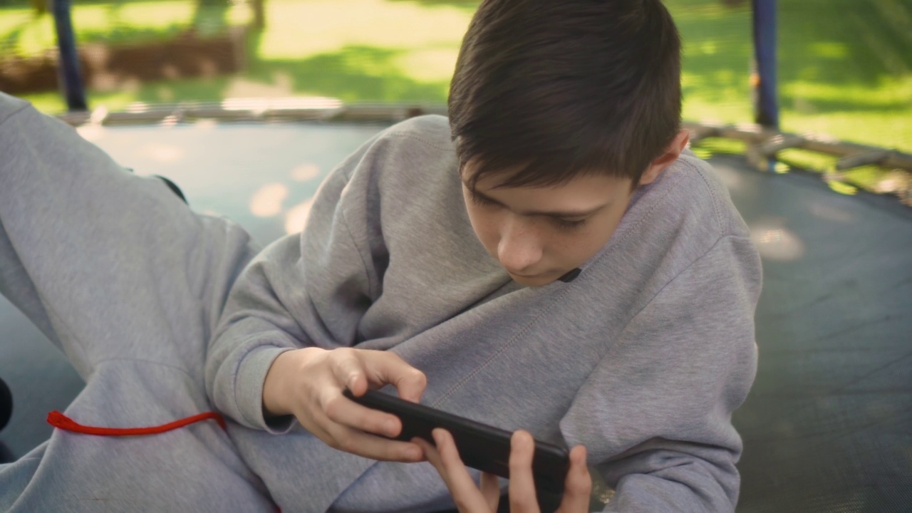 男孩在家院里躺在蹦床上玩手机游戏视频素材