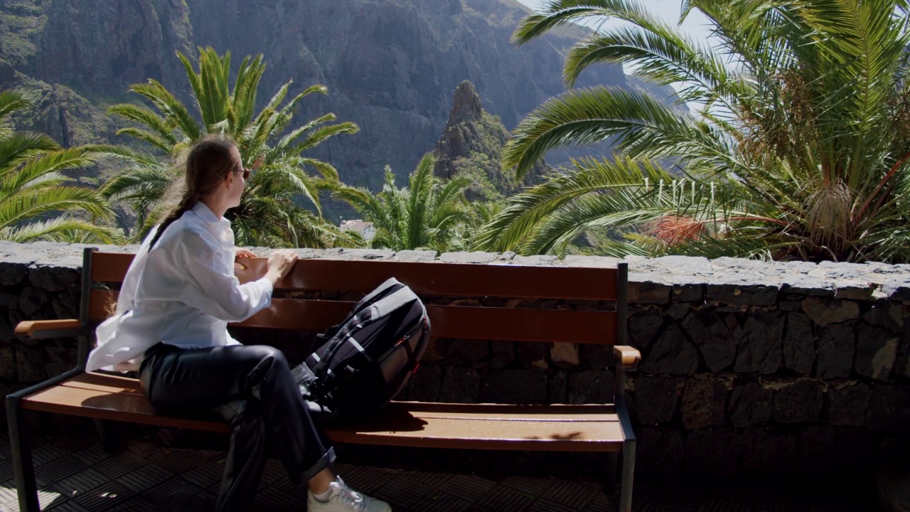马斯卡峡谷和特内里费岛的村庄。美丽的年轻女子背着双肩包坐在长椅上欣赏风景。慢动作视频素材