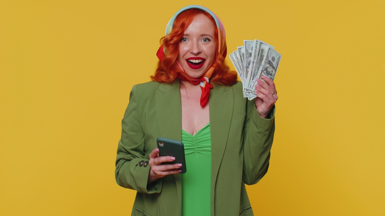 快乐的红发女子看着智能手机显示真诚的喜悦胜利，收到钱美元现金视频素材