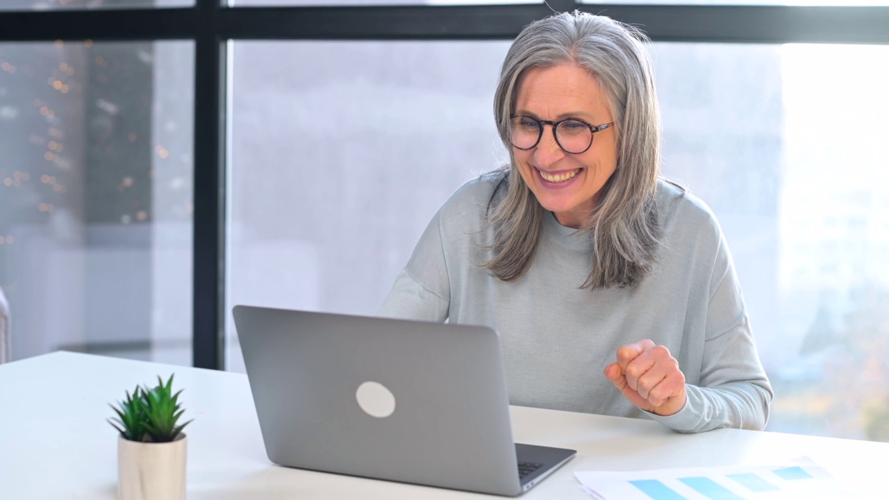 兴奋的灰白头发的高级妇女看着笔记本电脑屏幕坐在办公室的书桌视频素材