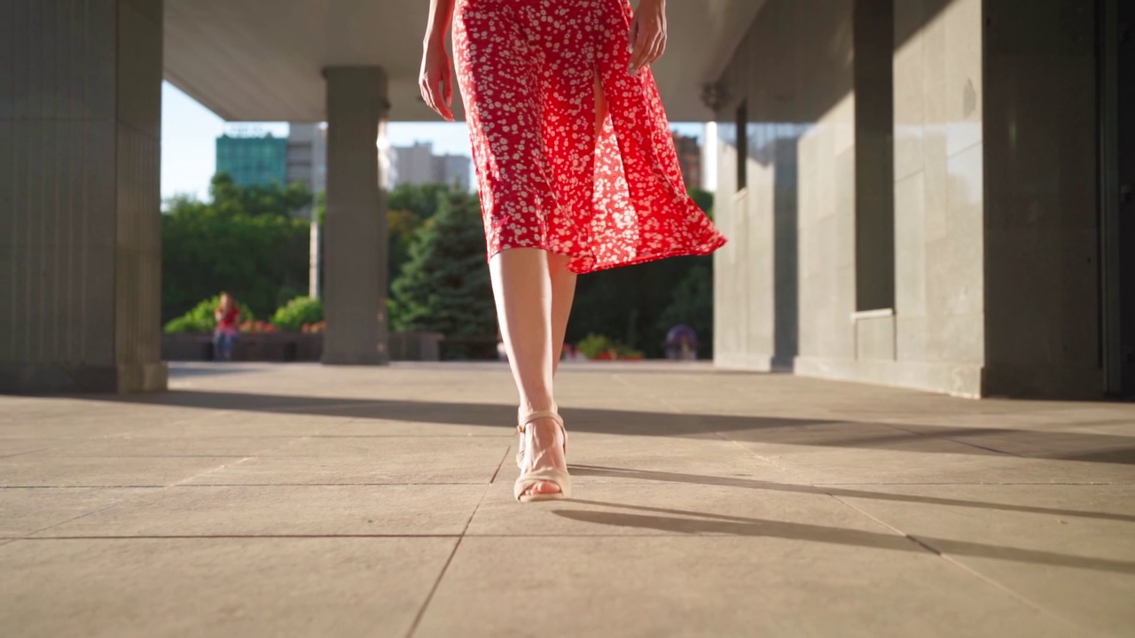穿着高跟鞋和红色连衣裙的苗条女性走在城市的阳光大道上。美丽的女孩漫步在夏日的市中心。认不出来的人。慢动作视频素材