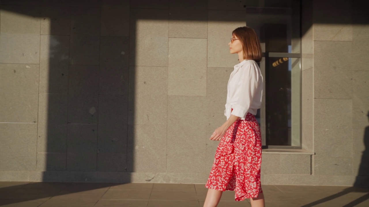 美丽苗条的女人在一个明亮的阳光明媚的日子里，戴着太阳镜和时尚的红裙子和白衬衫走在夏天的城市街道上。生活方式，女性华丽的脸户外。慢动作视频素材