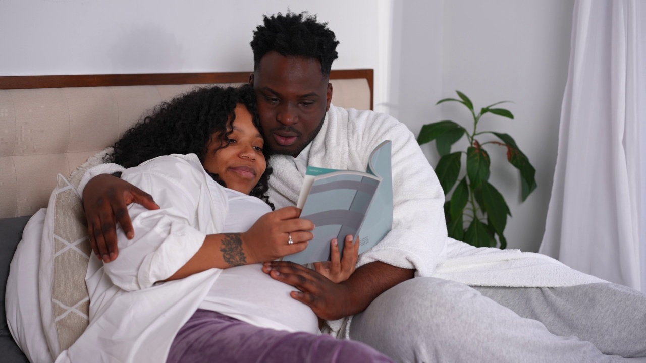 怀孕的非洲裔美国年轻女性躺在床上看书，男人在说话。一对轻松愉快的情侣周末在室内享受休闲时光。怀孕和家庭观念。视频素材
