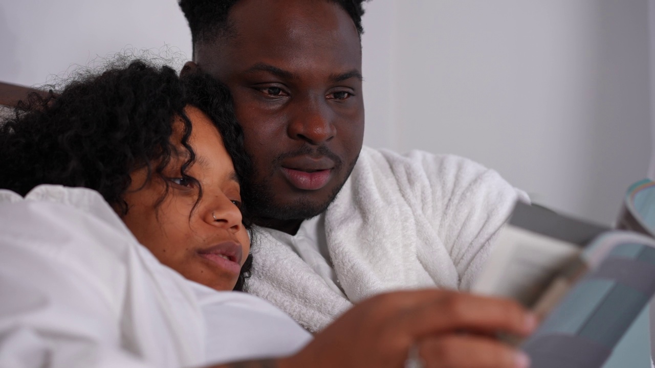 特写肖像快乐的非洲裔美国夫妇说话微笑读书躺在床上在家里。无忧无虑轻松的夫妻在卧室里一起享受爱好。爱和家庭。视频素材