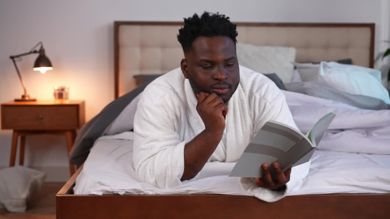 专心致志的非裔美国人快乐的男人躺在家里的床上看书。聪明、自信、放松的男人在卧室享受晚上的爱好的肖像。生活方式和快乐的概念。视频素材