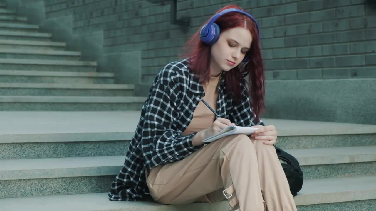 小女孩坐在楼梯上，戴着耳机，在笔记本上写东西视频素材