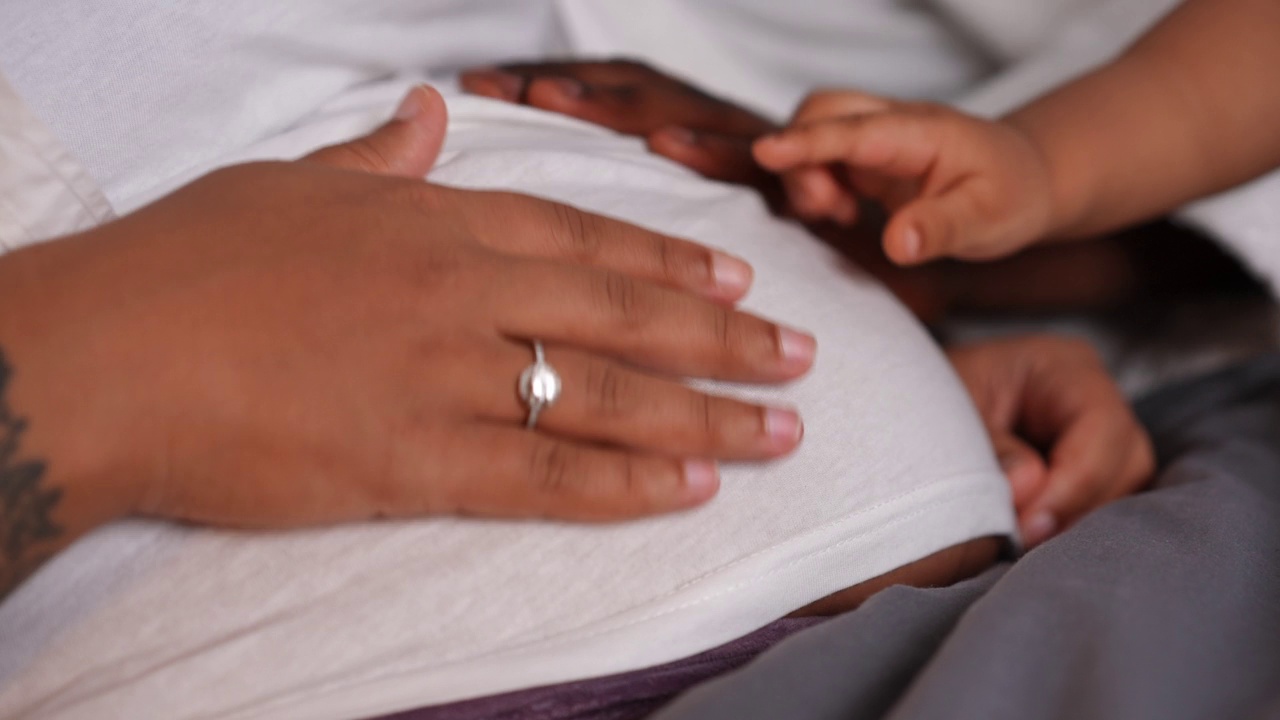 特写非洲裔美国年轻女性怀孕的肚子，用手抚摸肚子。面目全非的丈夫，妻子和小儿子抚摸着肚子躺在家里的床上。亲情的概念。视频素材