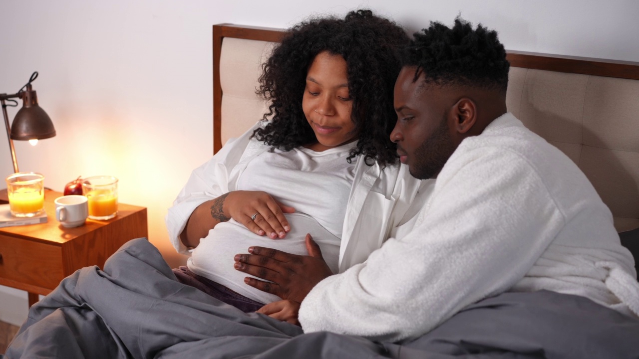 快乐的年轻非洲裔美国父亲听着说话，怀孕的肚子微笑的母亲可爱的蹒跚学步的孩子在卧室里奔跑。微笑的帅哥和美丽的女人躺在床上休息。视频素材