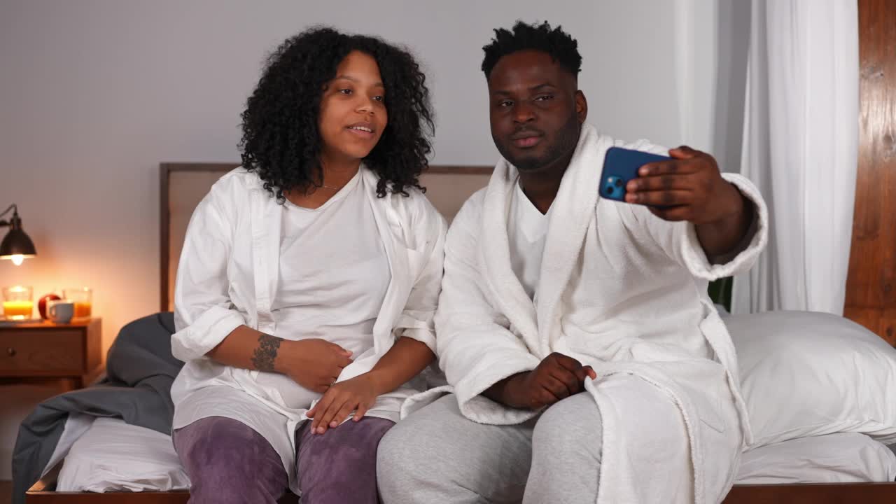 积极的非洲裔美国男子和孕妇在智能手机网络聊天挥手交谈。微笑着开心轻松的情侣在网上聊天，坐在家里的床上。便利和家庭生活方式。视频素材