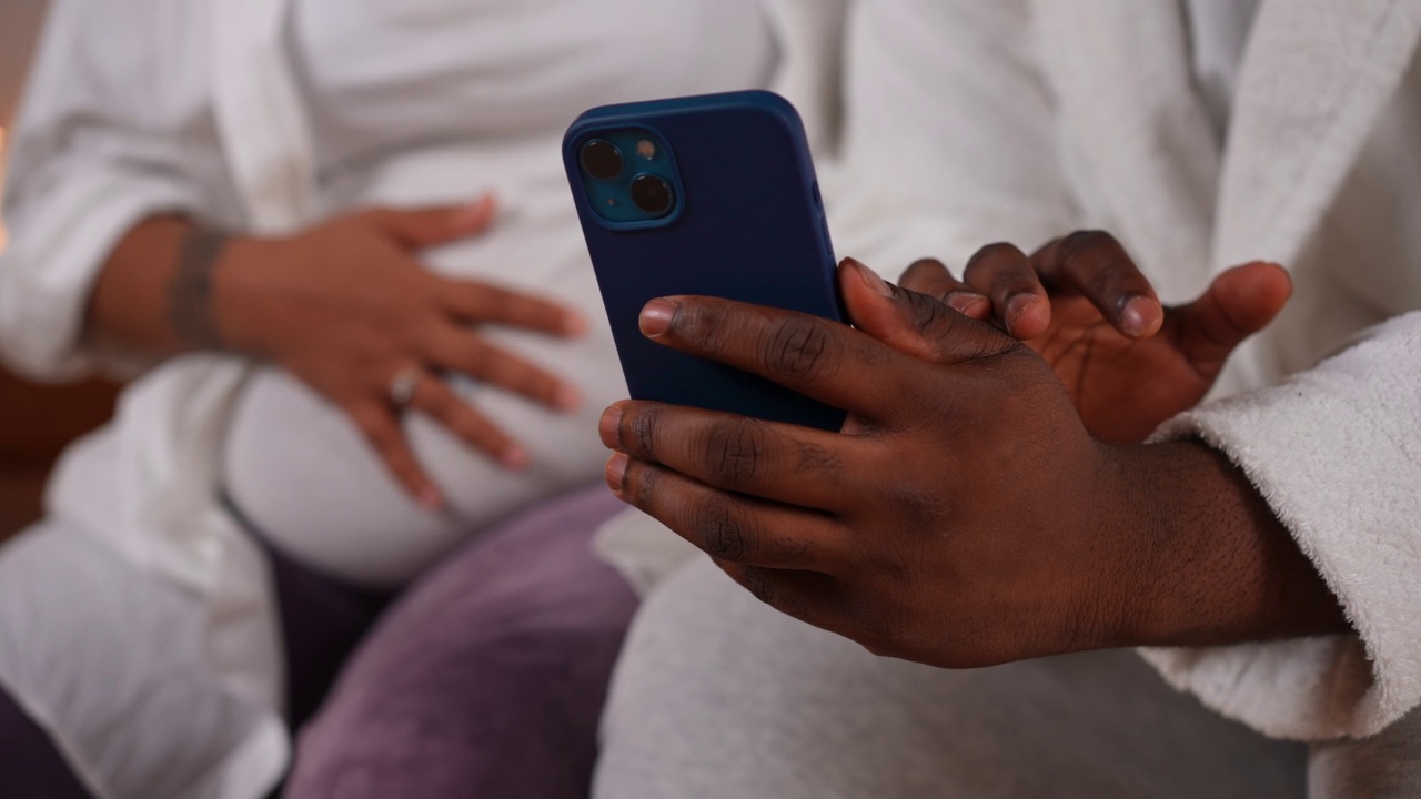 特写智能手机在年轻男子手中的手势与模糊的孕妇坐在背景。一对陌生的非洲裔美国夫妇在家里网上购物。视频素材