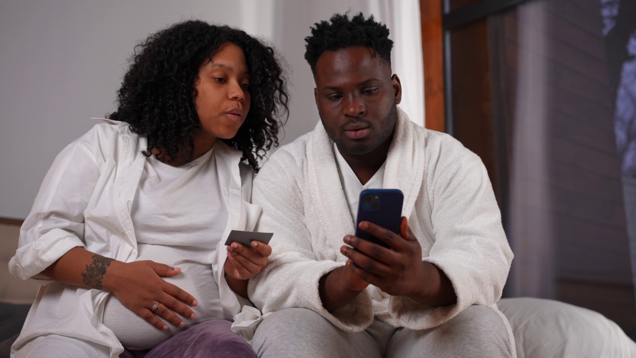 集中怀孕的非洲裔美国妇女告诉网站地址的男人在智能手机上网。专注的年轻夫妇坐在床上在家网上购物的肖像。视频素材