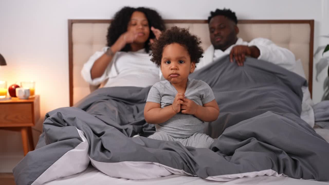 开朗可爱的非洲裔美国小男孩的肖像坐在床上，父母在背景交谈。快乐迷人的幼儿爬下离开卧室。休闲和幸福的概念。视频素材
