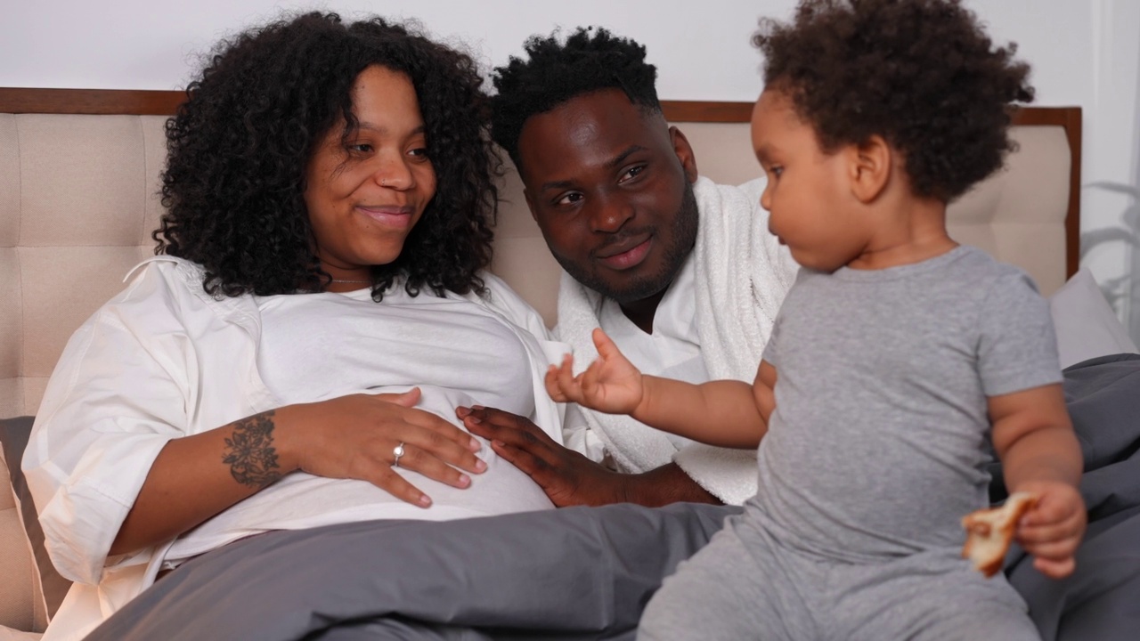 幸福的夫妻欣赏儿子触摸怀孕的肚子坐在床上的父母。微笑的非洲裔美国母亲和父亲享受周末休闲与可爱的蹒跚学步的小男孩。视频素材