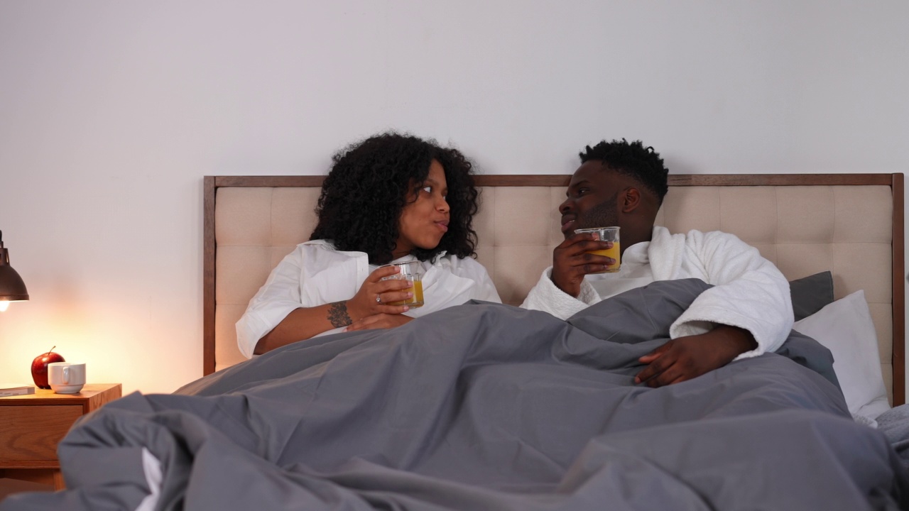 无忧无虑、快乐的非洲裔美国年轻夫妇早上在床上喝着橙汁聊天。积极怀孕的妻子和自信的丈夫分享在家里室内卧室聊天。视频素材