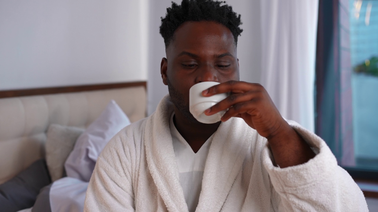 满意的英俊的非洲裔美国年轻人的肖像喝着早上的咖啡微笑着看着镜头。自信的男人在家里的卧室摆姿势，喝着美味的热饮。生活方式的概念。视频素材