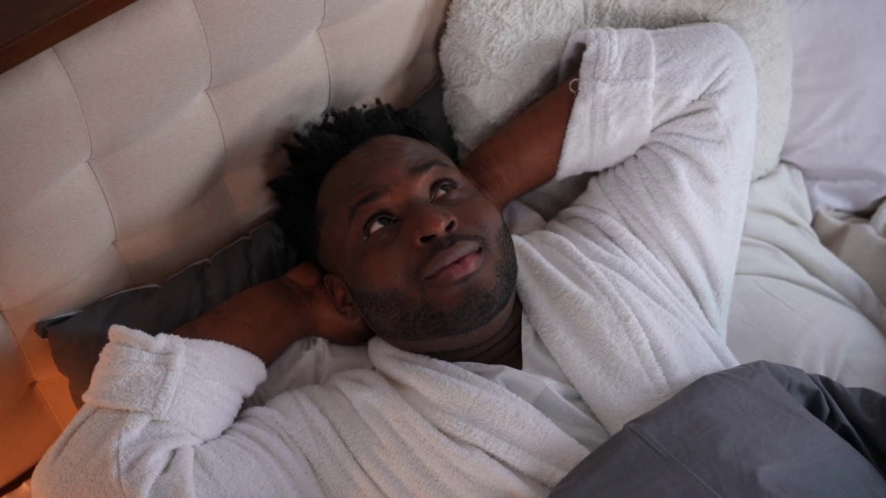 高角度视角肖像微笑快乐的非洲裔美国人躺在床上看向别处。轻松自信的帅哥穿着白色浴袍，在卧室的家里享受周末的休闲时光。视频素材