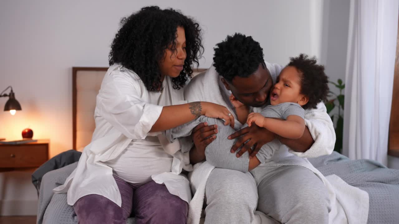 兴高采烈的非洲裔美国年轻父母给儿子挠痒痒，哄哭的孩子安静下来。快乐美丽的怀孕母亲和英俊的父亲与孩子在家里的卧室玩。视频素材