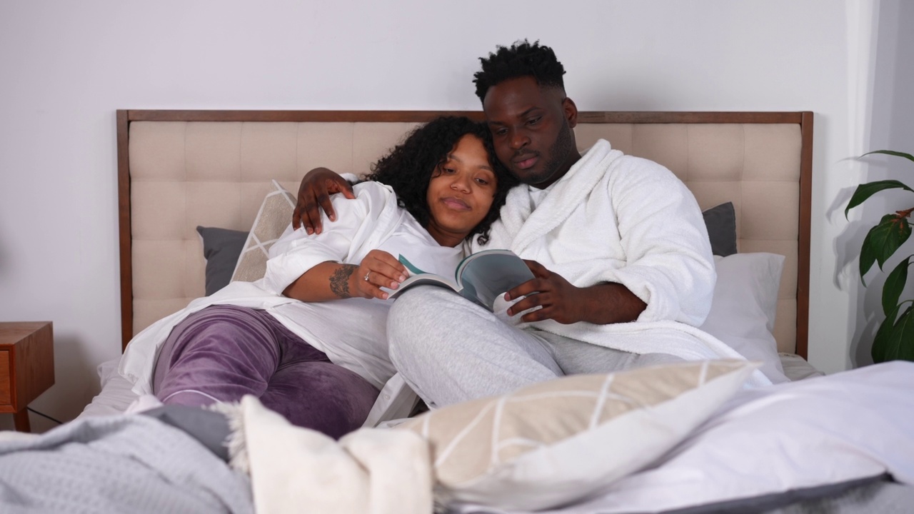 广角镜头爱丈夫亲吻妻子享受阅读与怀孕的配偶躺在床上。快乐自信的年轻非洲裔美国夫妇在家里的卧室里休息。视频素材
