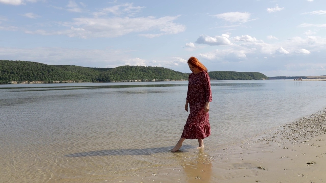 年轻的红头发，美丽的女人穿着红裙子，光着脚在山间湖泊的沙滩上画画视频素材