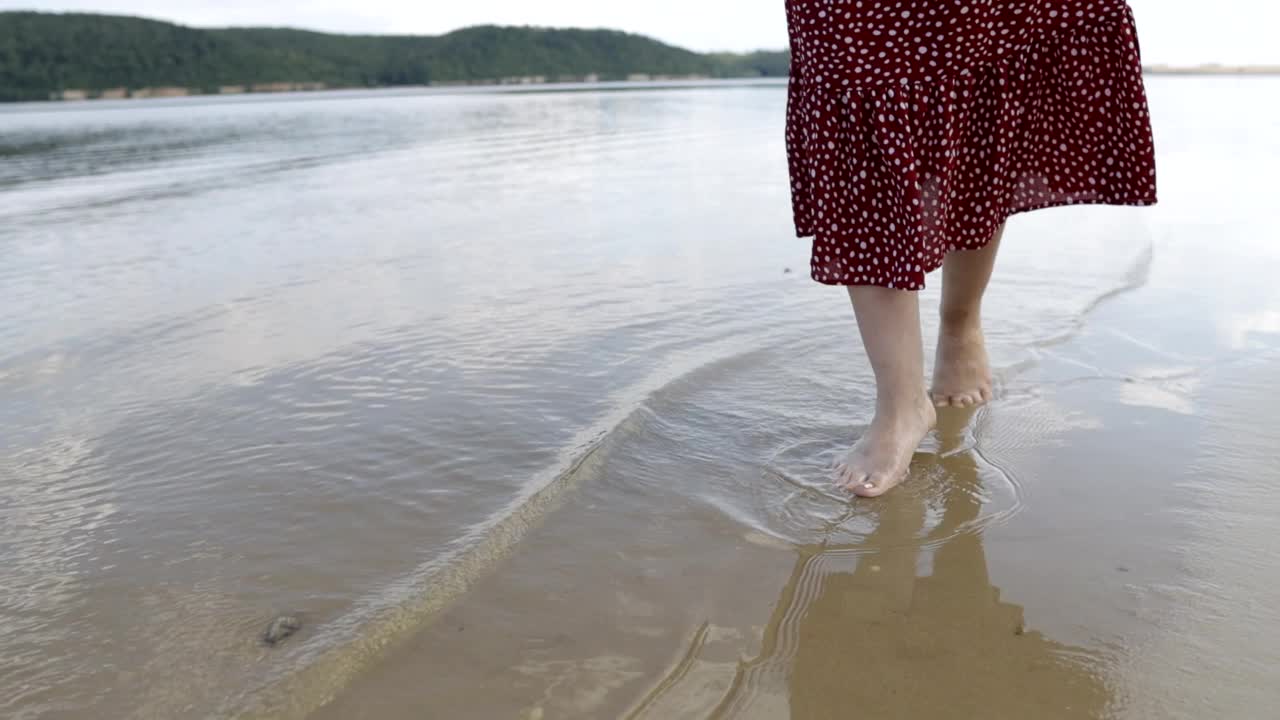 身着红色连衣裙的女性双腿沿着山间湖泊的沙滩奔跑，体现了与自然统一的理念视频素材