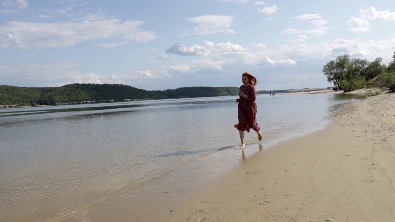 年轻的红头发、美丽的女人穿着红裙子沿着山中的沙滩跑视频素材