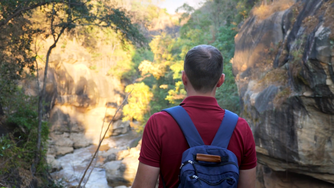 一名男性游客背着双肩包，在日落时分徒步游览自然公园。少年远足户外，发现山间河流。夏日冒险，假期自由理念。在野外远足。后视图视频素材