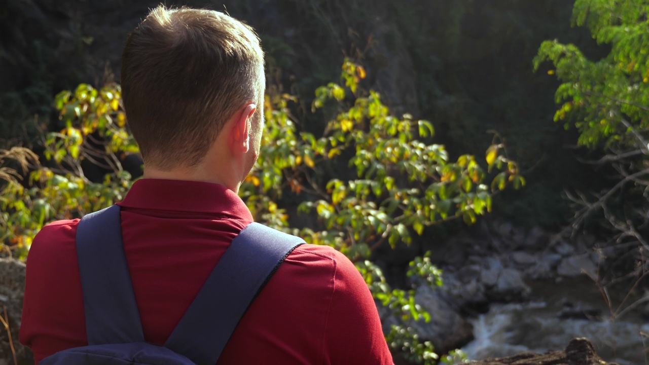 背包旅行者站在山边的背影，在日落的峡谷中欣赏自然的河景。森林里的男性徒步旅行者，风景优美。视频素材
