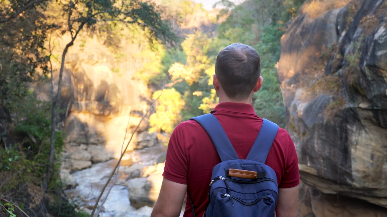 年轻男子背着双肩包在自然公园徒步旅行。男游客户外远足，发现山间河流。在野外远足。后视图。日落。夏日冒险，假期自由理念视频素材