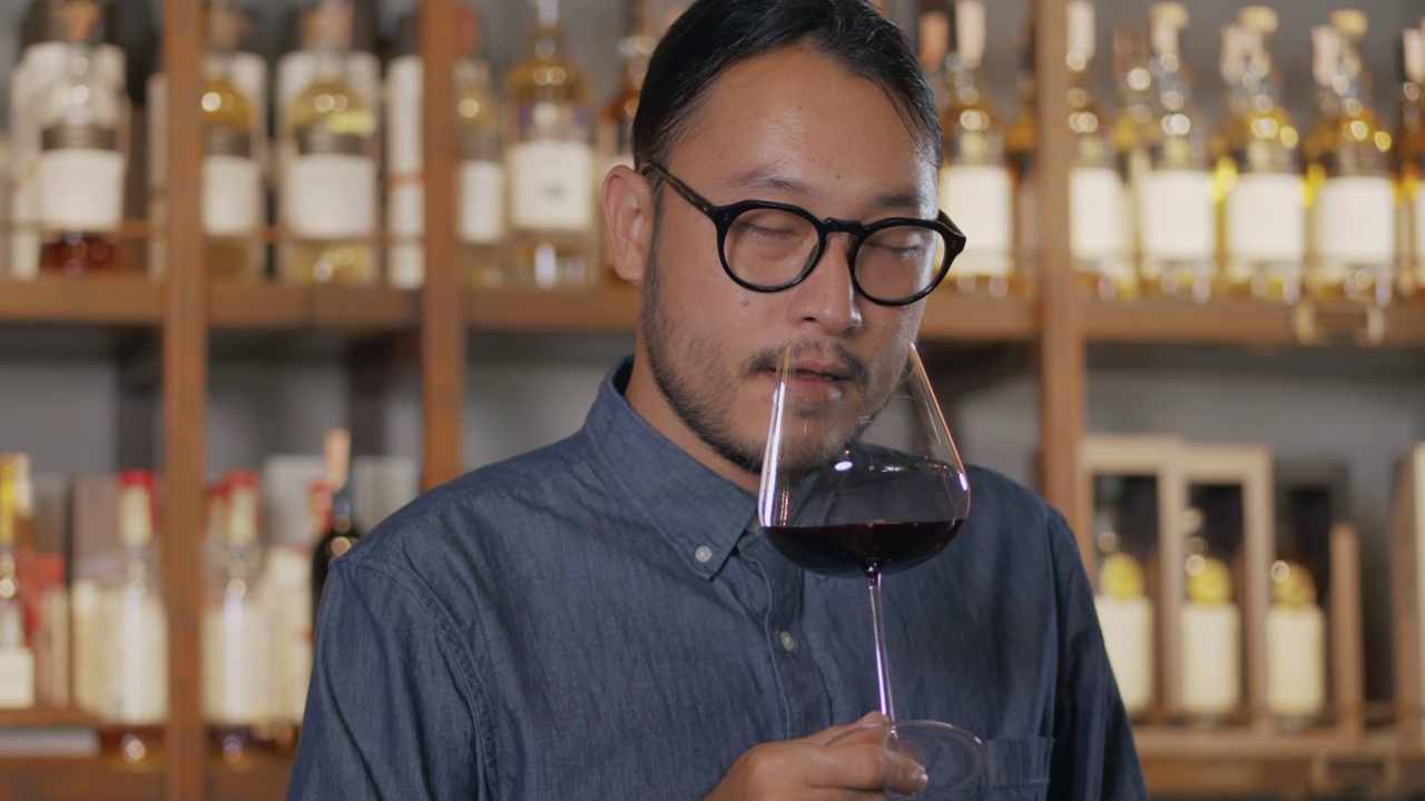 年轻的亚洲侍酒师在品尝新进口的葡萄酒时，微笑着。戴眼镜系领带的男子在酒店的餐厅酒吧品酒。视频素材