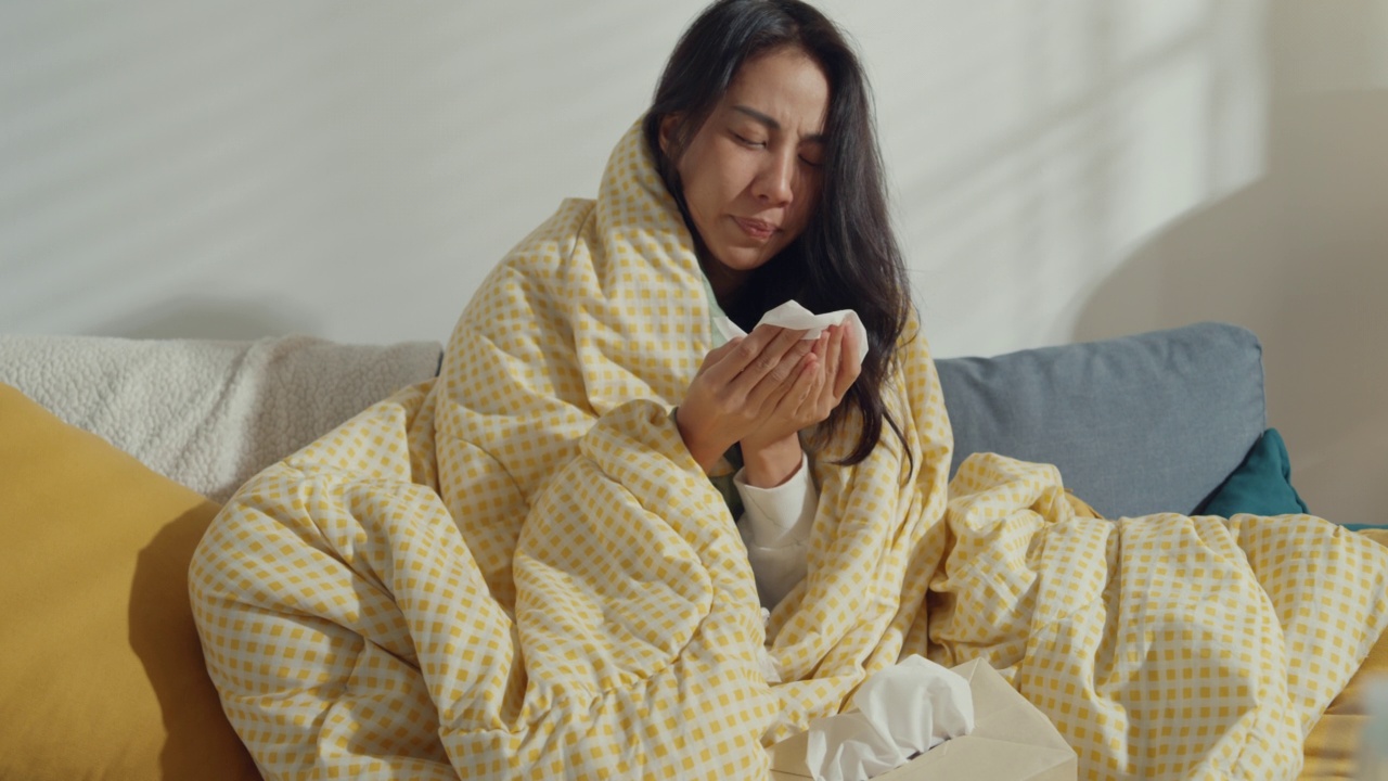 生病的年轻亚洲妇女擤鼻涕和打喷嚏用纸巾坐在毯子下的沙发在家里的客厅。视频素材