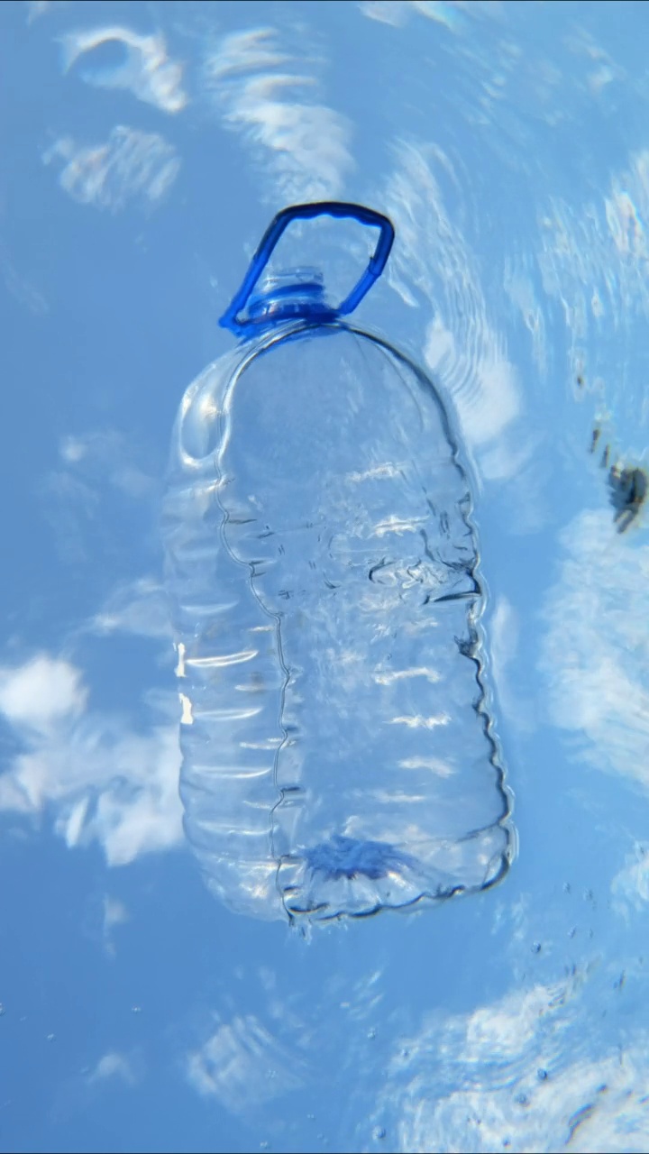 大塑料瓶漂浮在水面上，背景是蓝天白云。有柄的桶在海浪中漂流。海洋塑料污染。水下拍摄。垂直视频视频下载