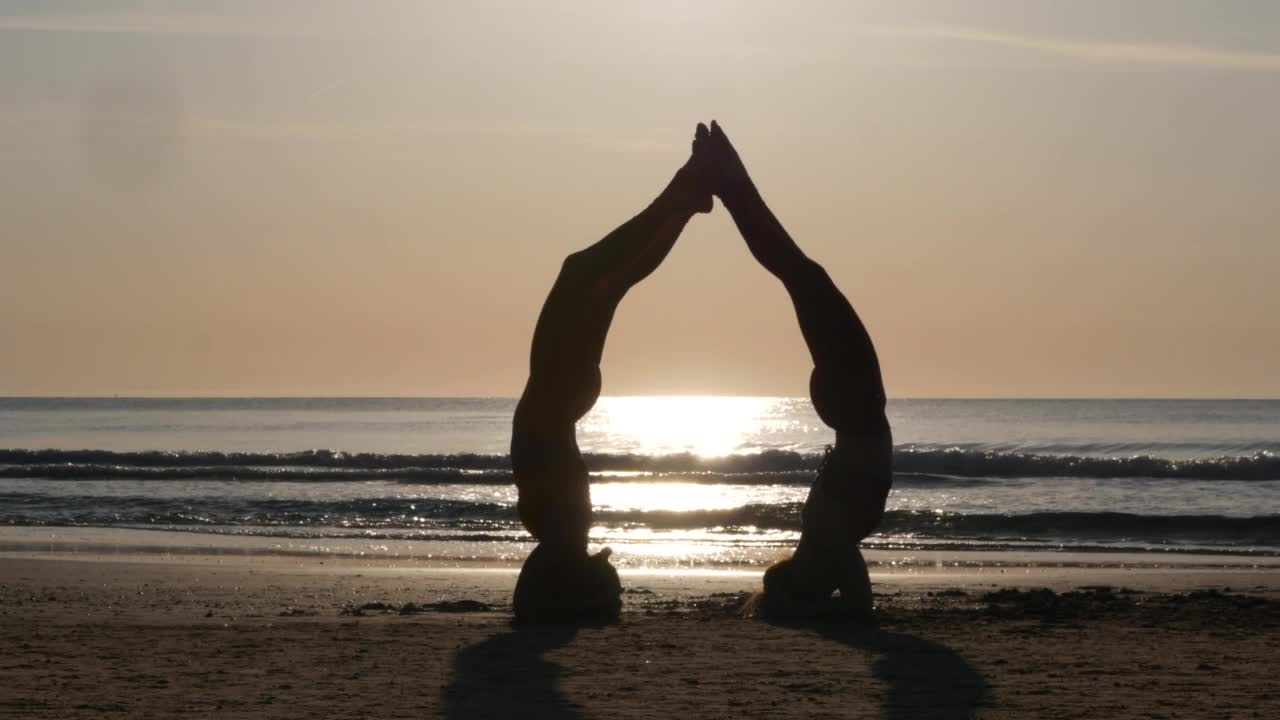 两名女子在日出时在海滩上做瑜伽，头倒立姿势视频素材