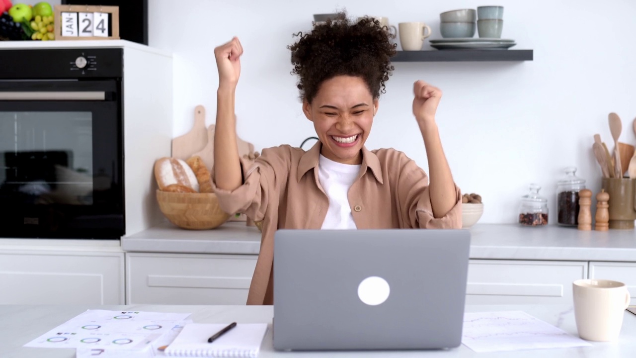 兴奋快乐的非裔美国卷发女孩、经理、股票经纪人、设计师，惊喜地看着笔记本电脑屏幕，看到意想不到的消息，为项目的成功而高兴，微笑视频素材