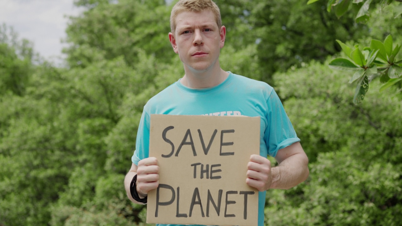 肖像，正面视图:白人青年男子志愿者拿着纸板认真为拯救地球的运动，不使用塑料，拯救绿色环境，靠近森林视频下载