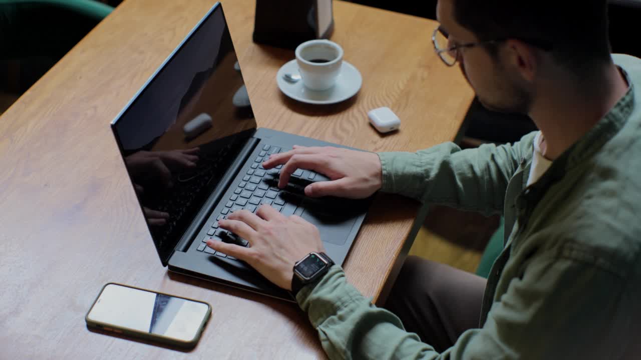 俯视图是一个穿着休闲服的男人坐在桌子旁，在笔记本电脑上打字。学生在咖啡厅用咖啡杯学习。人与科技的概念。视频素材