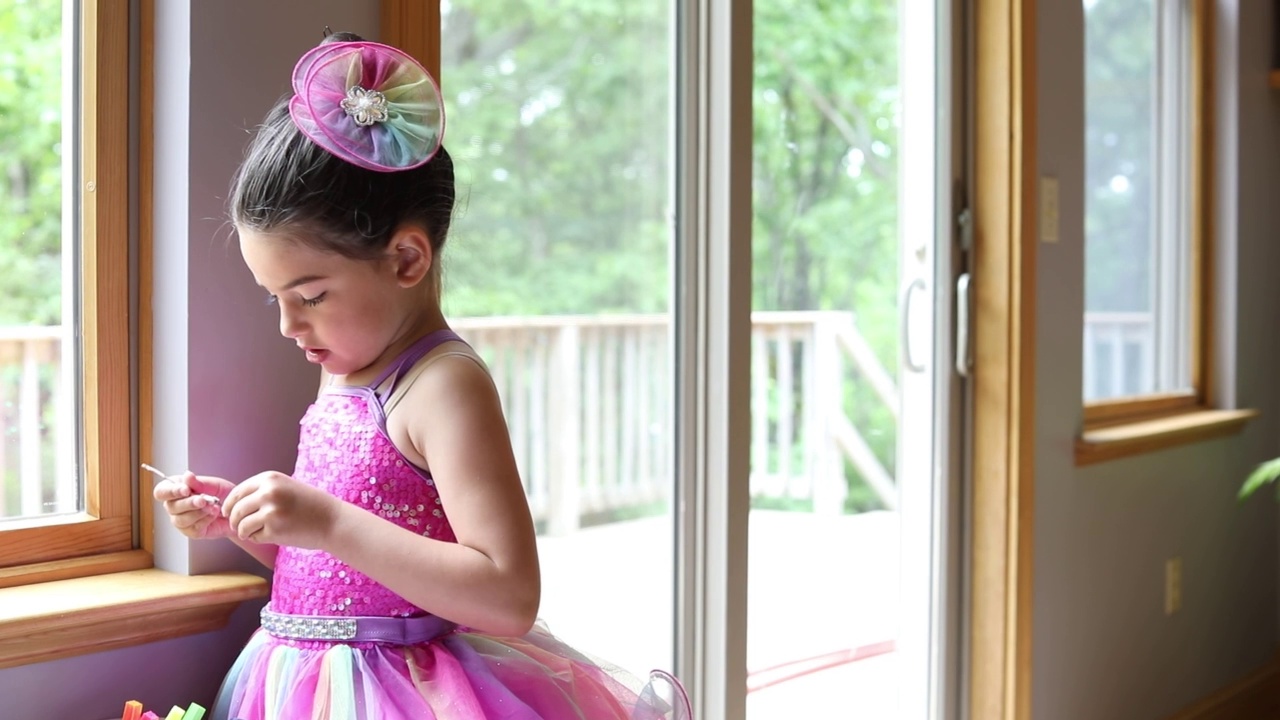 5岁的女孩站在窗户的灯光下，穿着粉红色的亮片舞蹈服装，专注地看着她精致地握在手里的一根柳枝，然后把它举到镜头前。视频下载