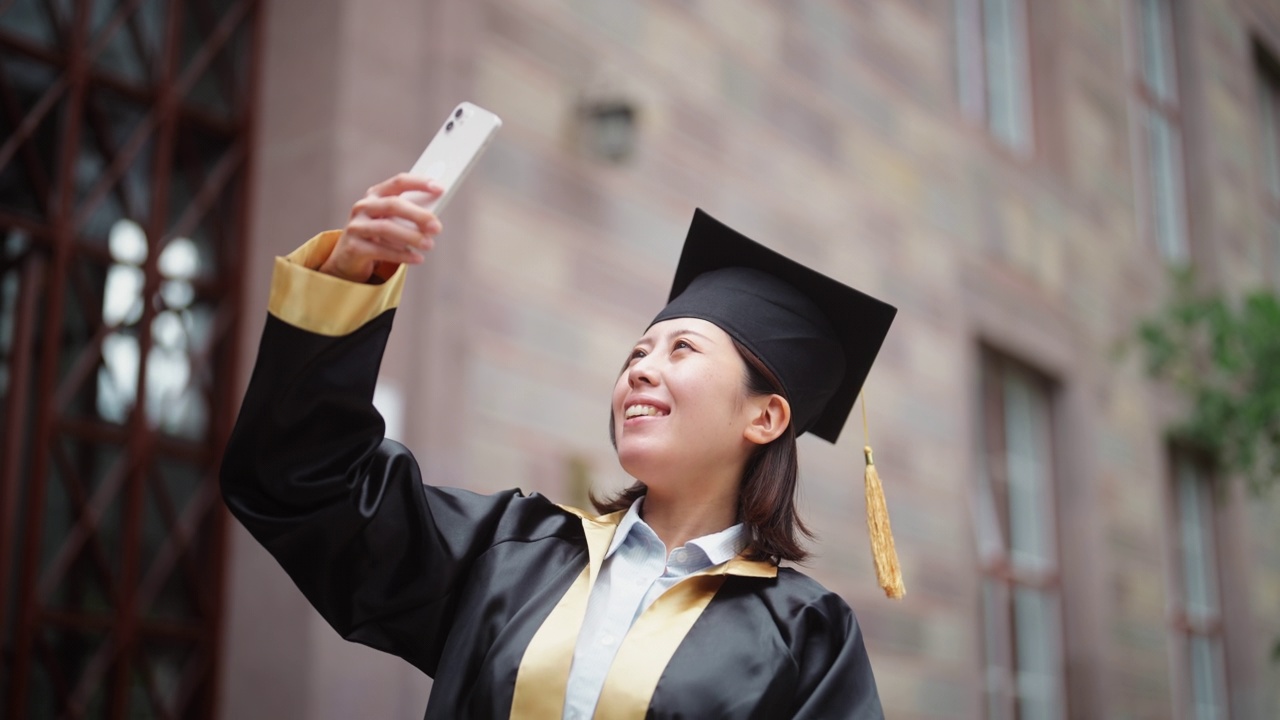 身着毕业礼服的亚裔女大学生在校园里用智能手机自拍视频下载