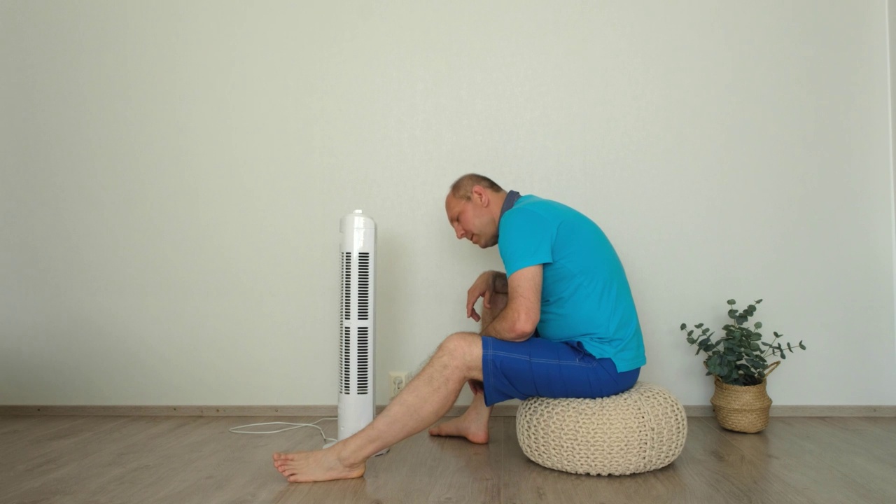 热，一名白人男子坐在风扇旁，躲避夏天的炎热。视频素材