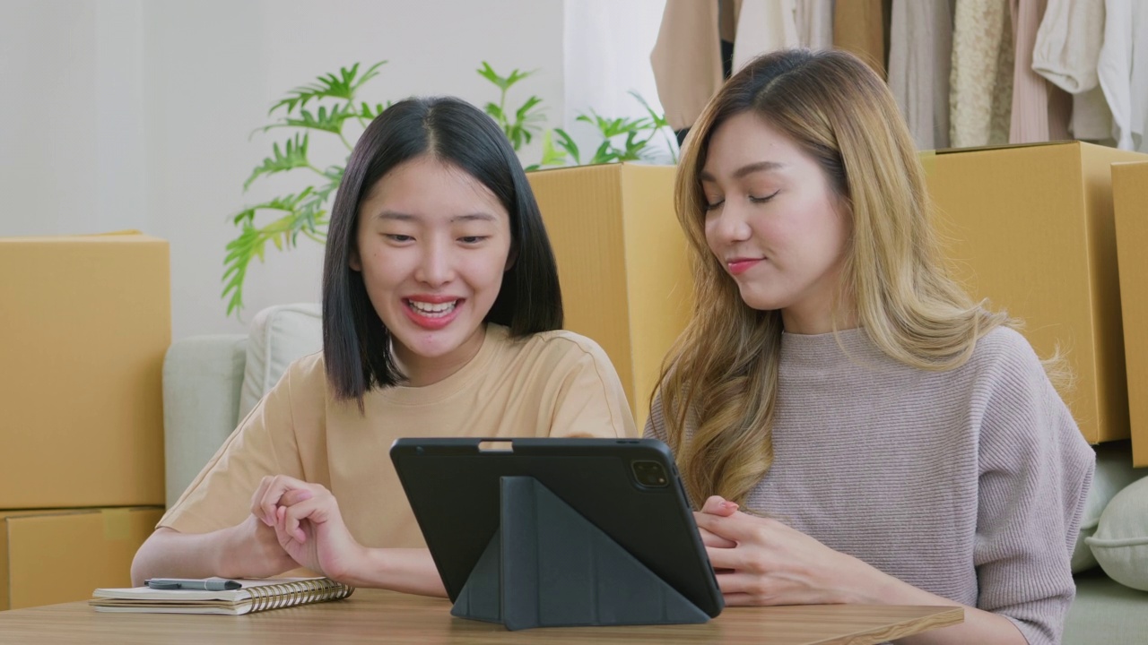 年轻的亚洲夫妇在卖衣服后，开始计算收入和支出。几个女人坐在一张小桌子旁，在家用平板电脑做会计。视频下载