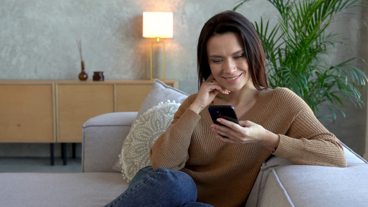 快乐愉快的女人放松在舒适的沙发上，手里拿着智能手机视频下载