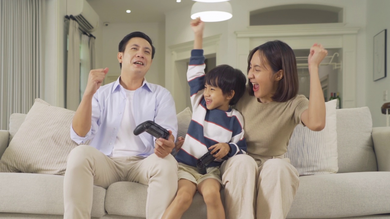幸福微笑的亚洲家庭的肖像生活在一起的家里，玩游戏与控制器，坐在沙发上的家庭关系。父亲、母亲和儿子的爱。人们的生活方式。视频下载